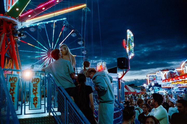 A couple stands on a ramp at a fair at night, with a Ferris wheel and other rides in the background.