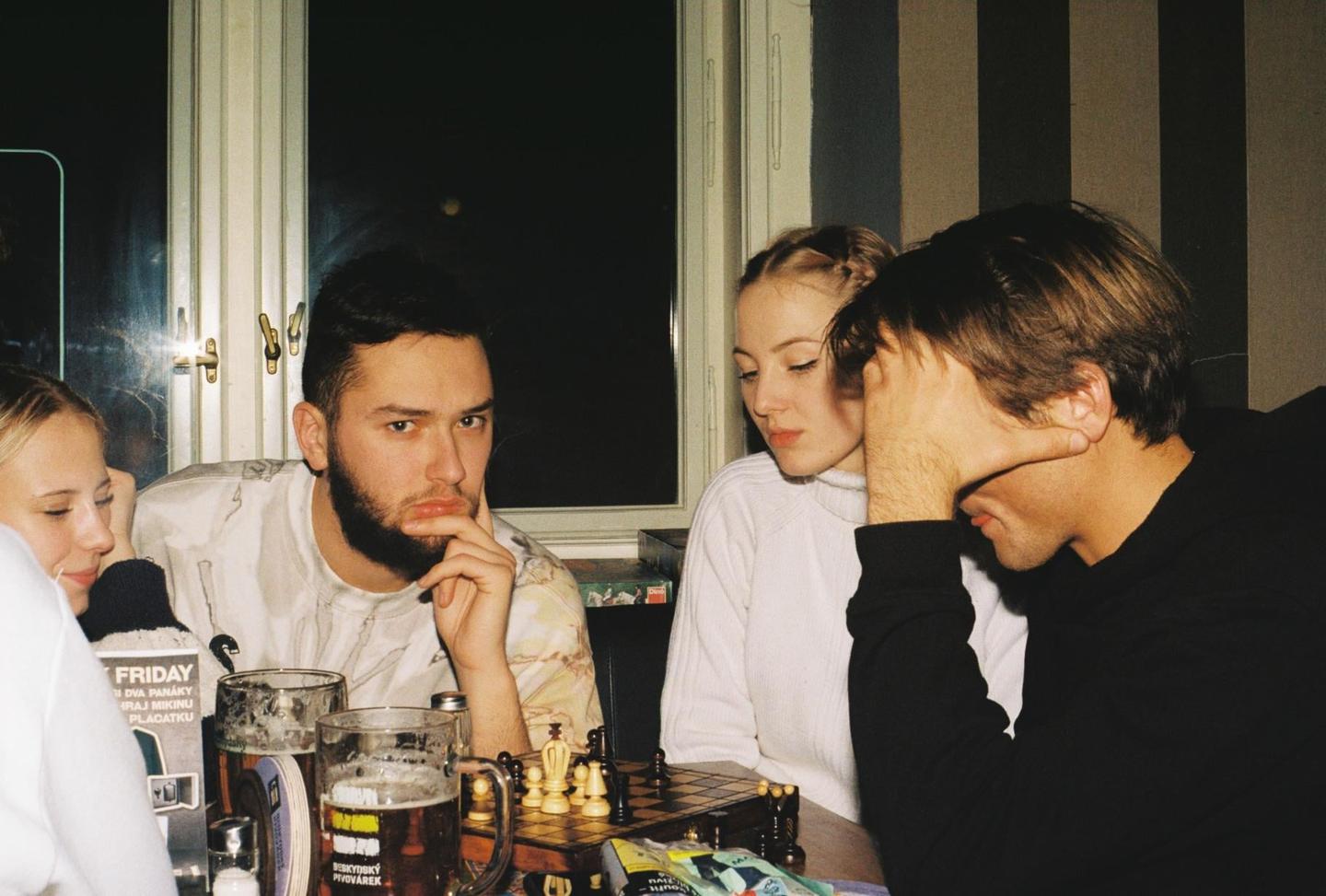 Three young adults sit around a table with a chessboard and beer mugs, with one man looking directly at the viewer.