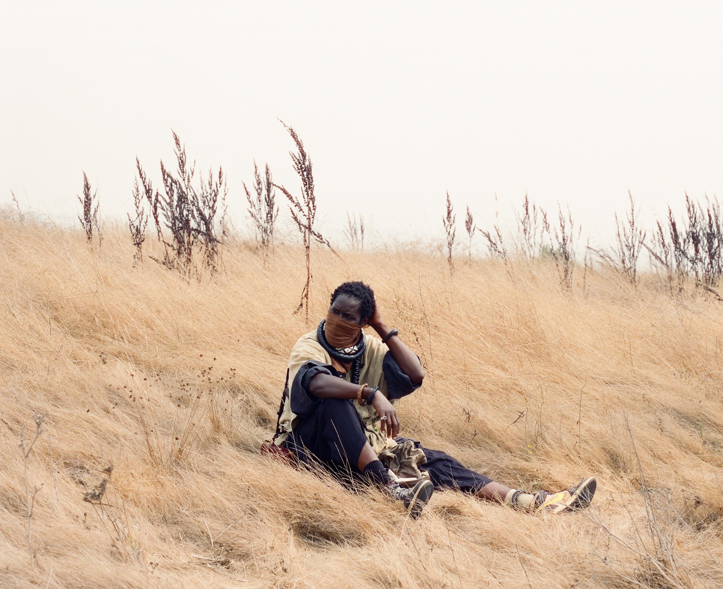 A man wearing a face covering and traditional clothing sits in a field of dry grass.