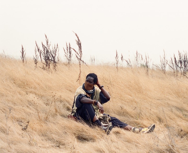 A man wearing a face covering and traditional clothing sits in a field of dry grass.