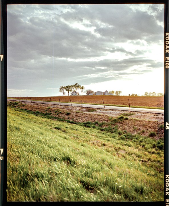 A rural landscape shows a green grassy slope in the foreground, a dirt road with a fence, and a farm with a house, silos, and trees under a cloudy sky.