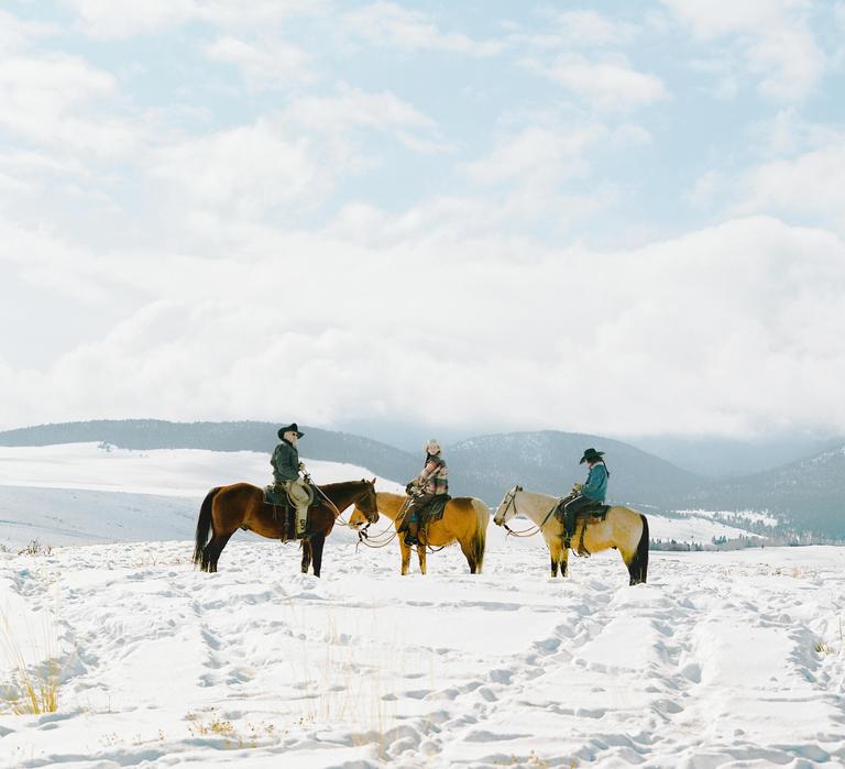 Three people on horseback are in a snowy field with mountains in the background.