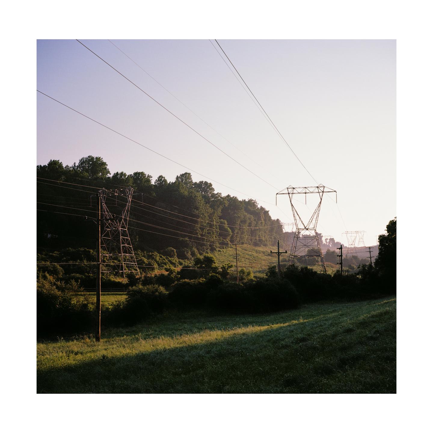 Power lines stretch across a hilly landscape with trees and a grassy field.