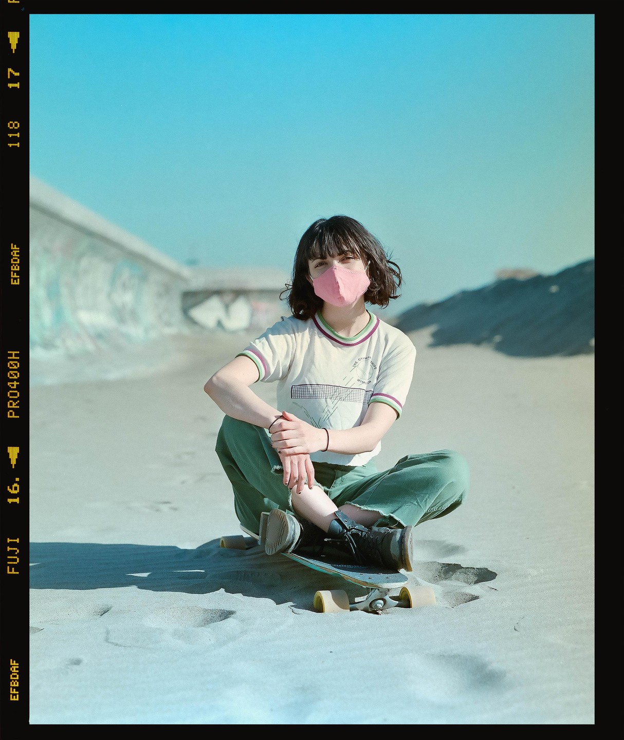 A woman wearing a pink mask sits on a skateboard in the sand.
