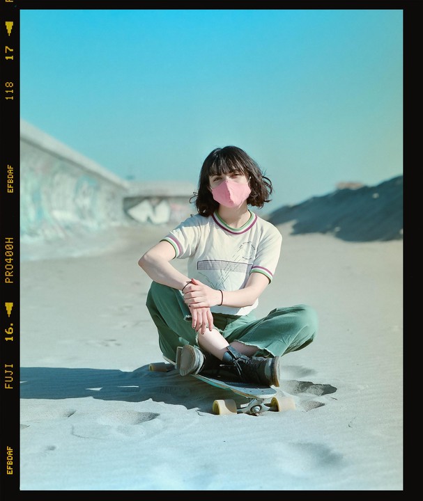 A woman wearing a pink mask sits on a skateboard in the sand.