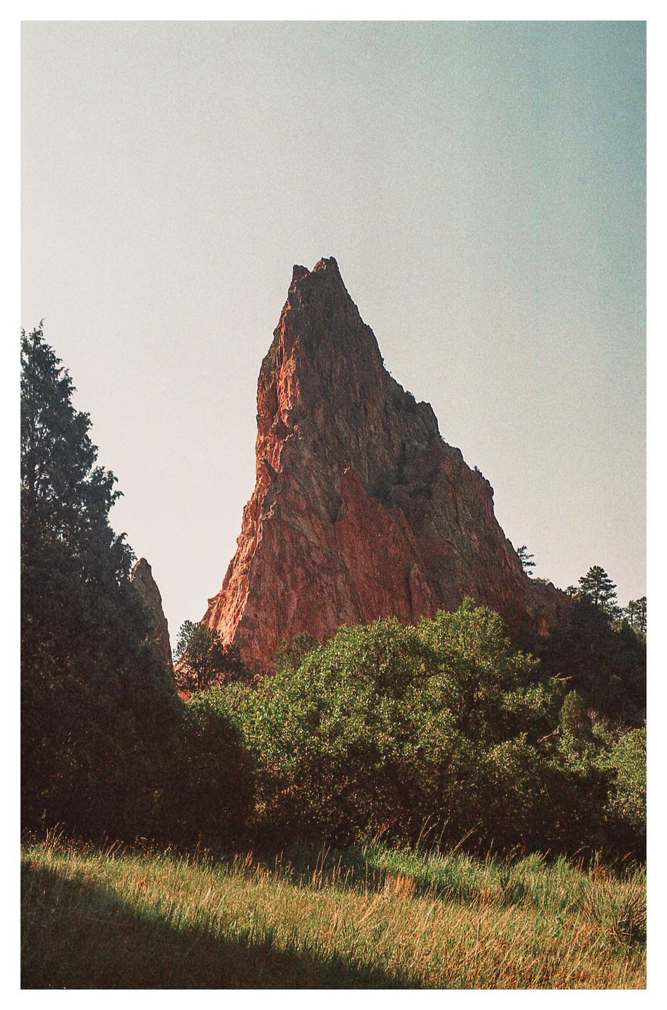 A large red rock formation rises above green trees and a grassy field under a clear sky.