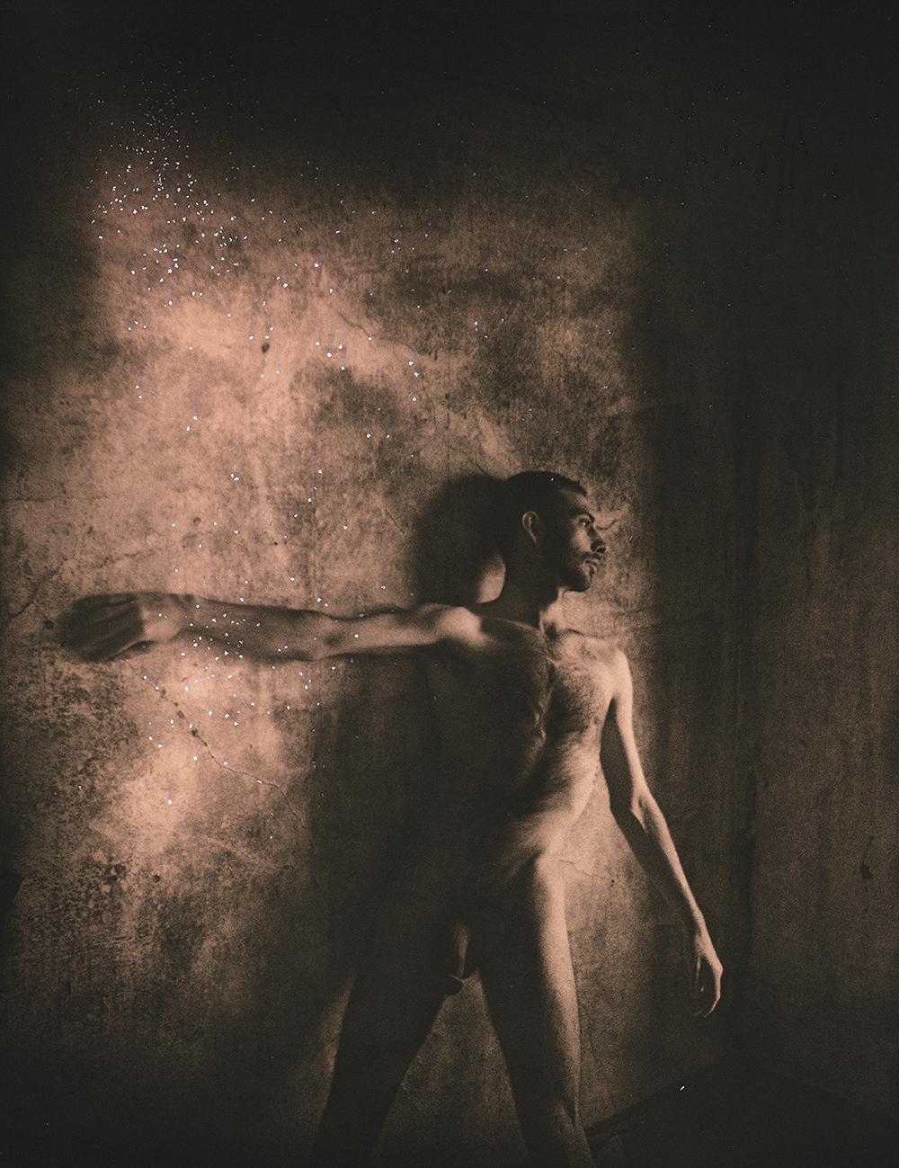 A man stands nude with his arm outstretched against a textured wall with sparkling dust.