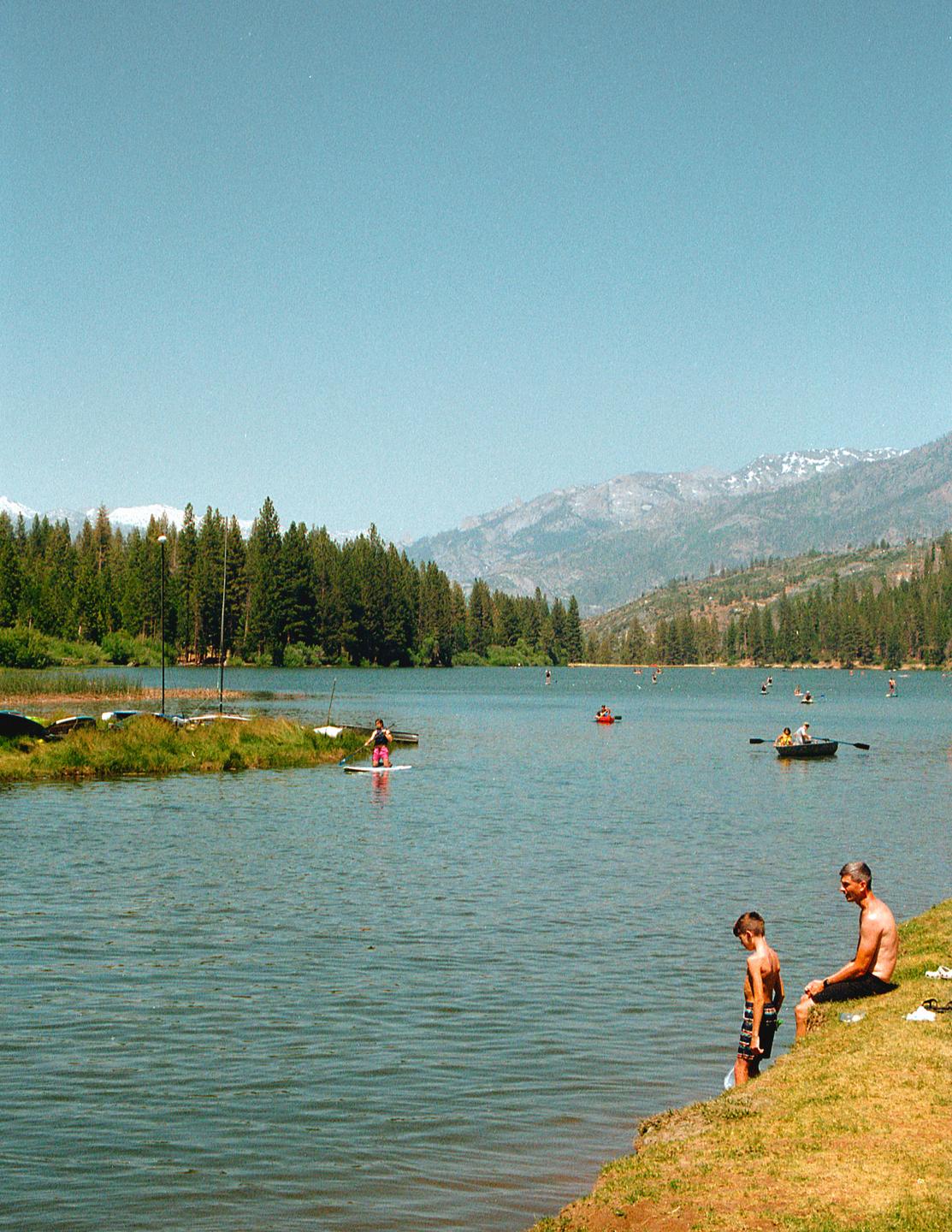 A man and a boy sit by the edge of a lake with mountains and a forest in the background.