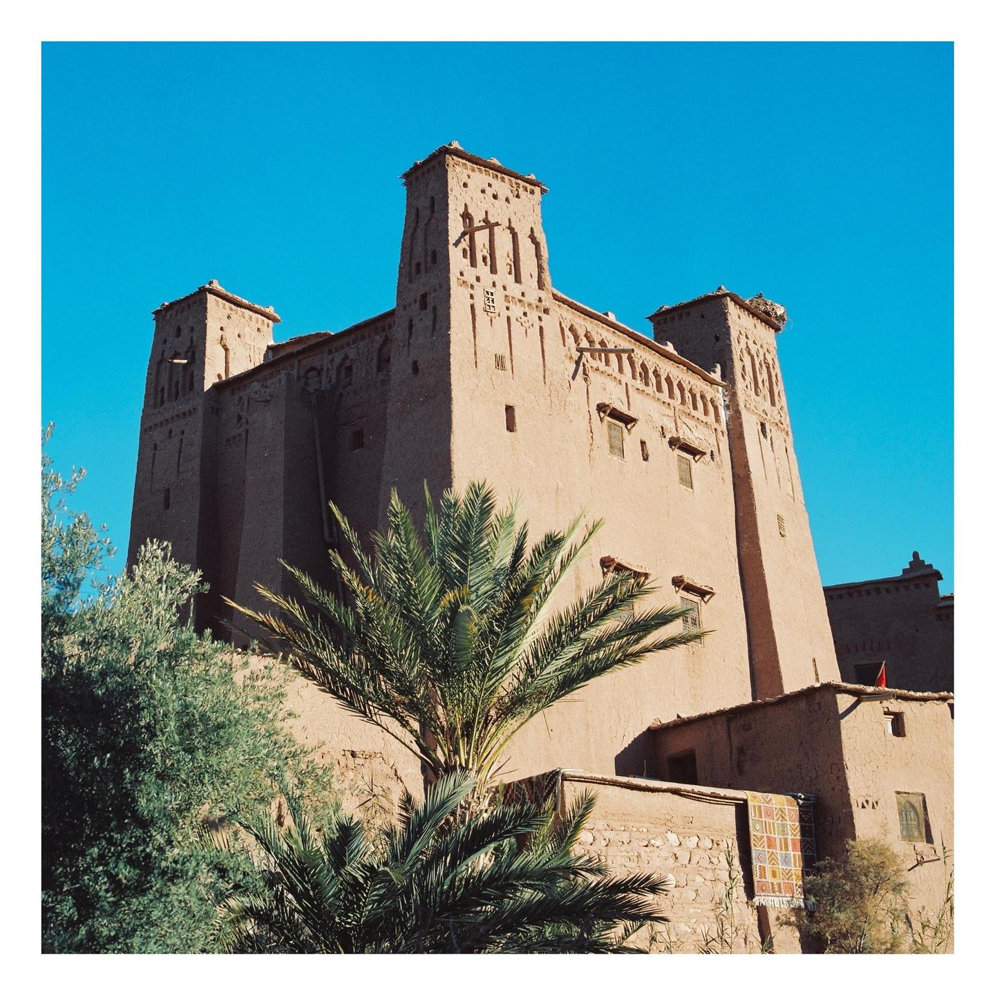 A tall, ancient mud-brick building with towers stands against a clear blue sky, with palm trees in the foreground.