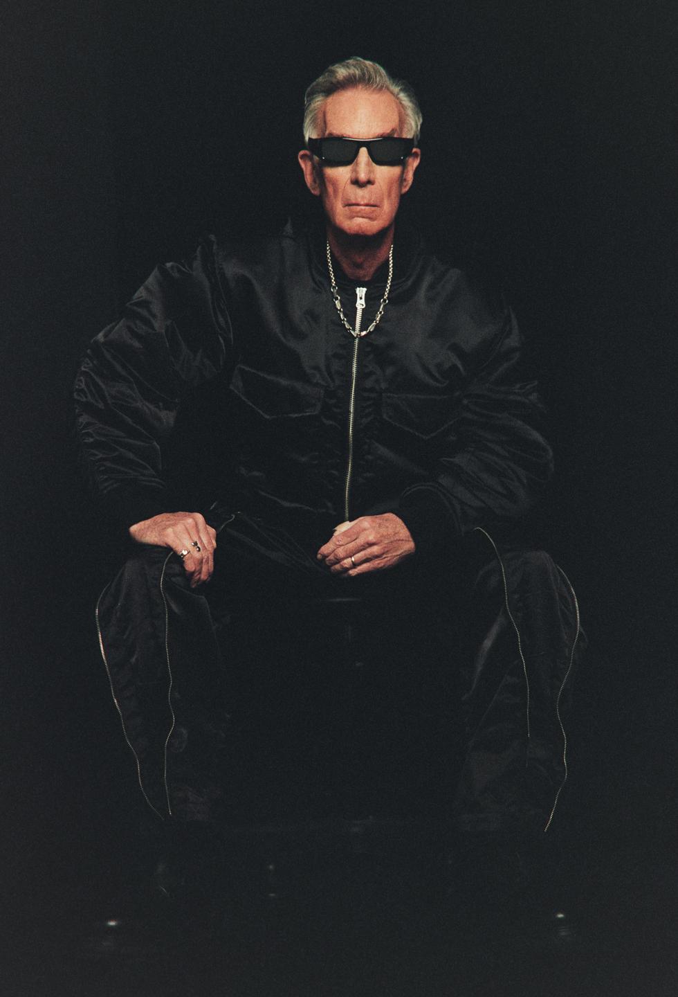 A man wearing sunglasses and a black jacket is crouching in a dark setting.