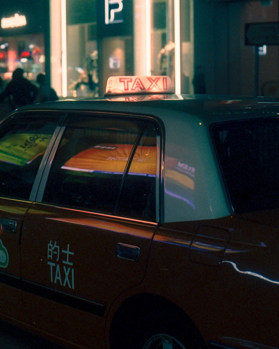A taxi is parked on a city street at night, with reflections of neon lights visible in its windows.