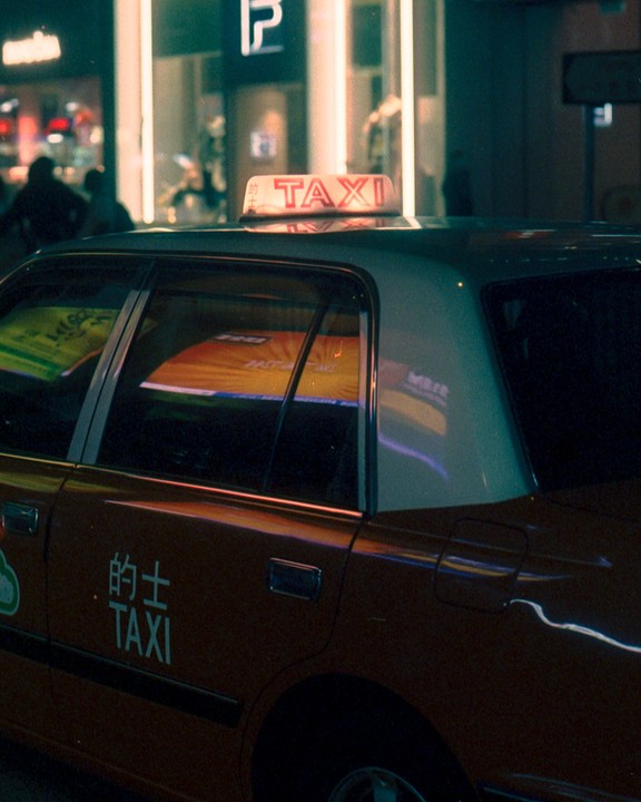 A taxi is parked on a city street at night, with reflections of neon lights visible in its windows.