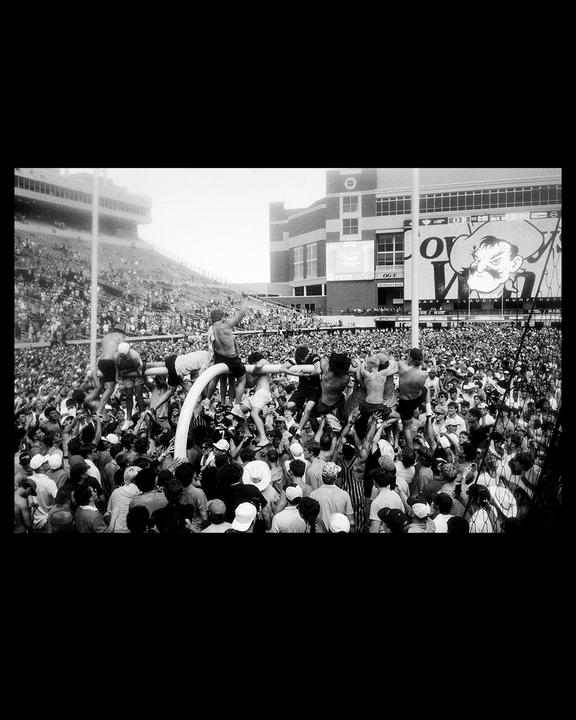 A large crowd of people is gathered on a sports field, with some individuals climbing on a goalpost.