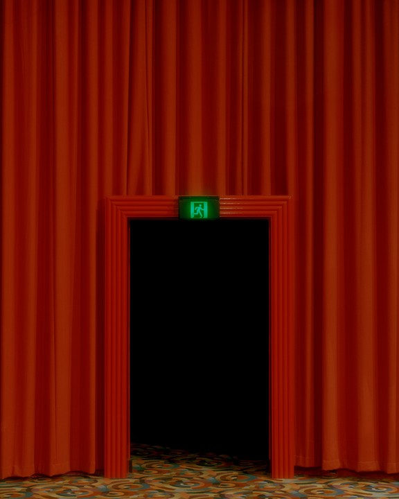 A green exit sign is visible above a dark doorway framed by red curtains and a patterned carpet.