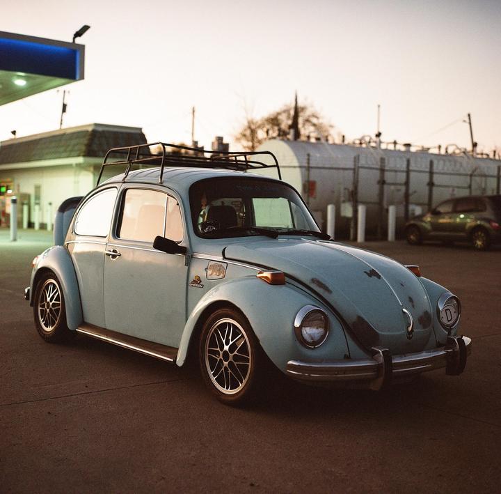 A light blue Volkswagen Beetle with a roof rack is parked at a gas station at dusk.