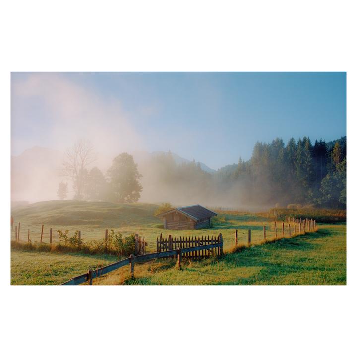 A small wooden hut sits in a foggy, grassy field with a wooden fence and trees in the background.