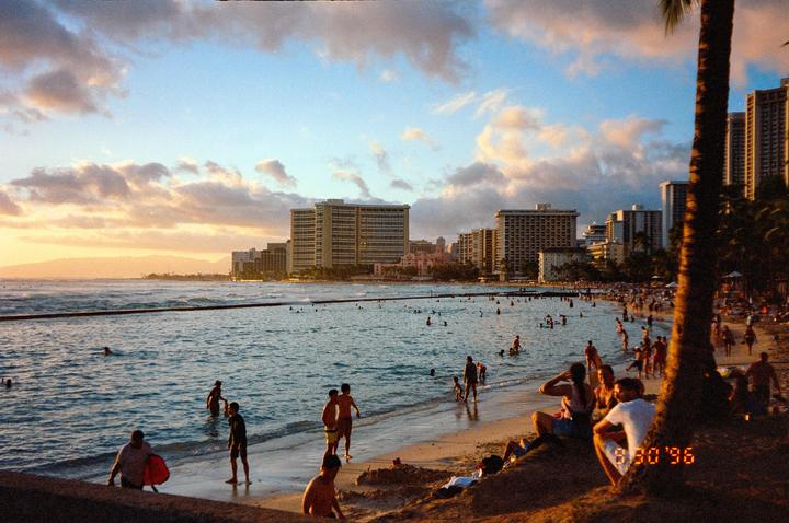A beach is crowded with people swimming and relaxing on the sand as the sun sets behind distant mountains and city buildings.