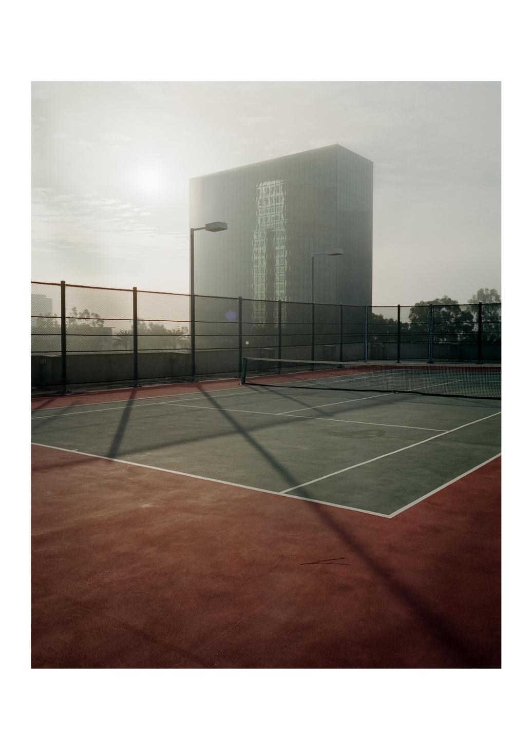 A tennis court with a net and fence is in the foreground, with a large building and hazy sky in the background.