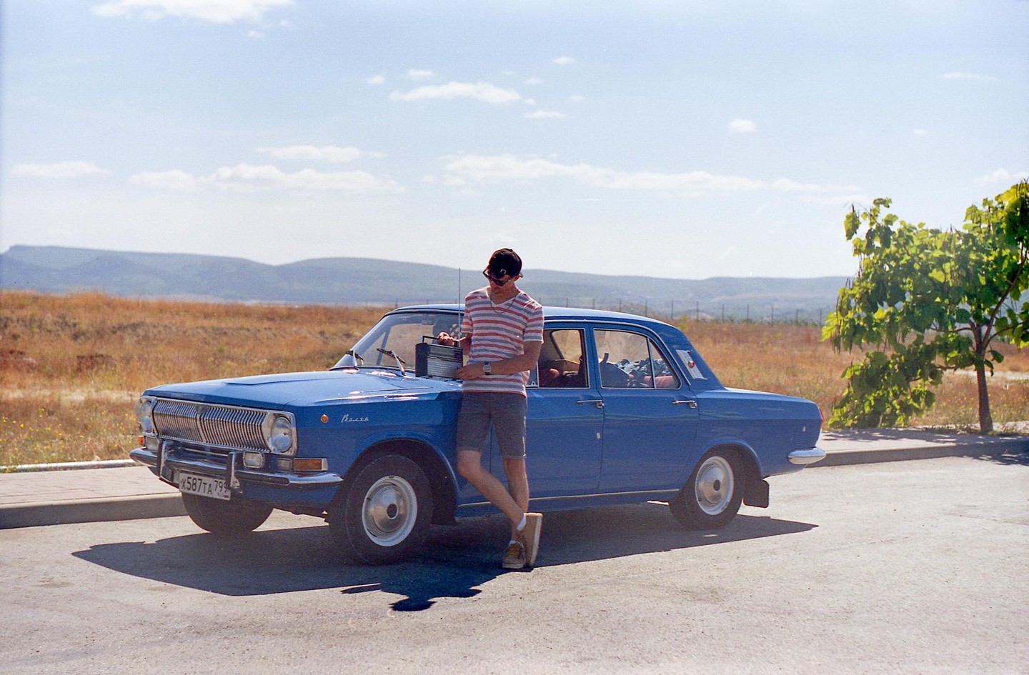 A man in a striped shirt stands next to a blue car with a radio on the hood, with a tree and hills in the background.
