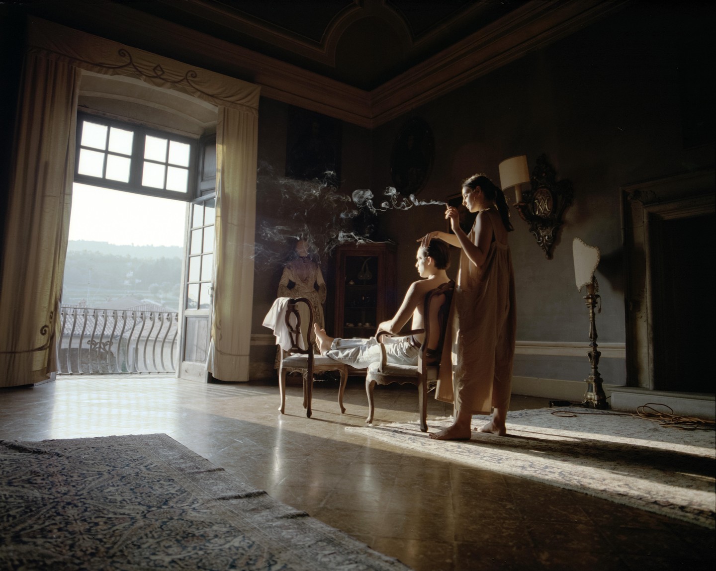 A woman styles another woman's hair in a sunlit room with an open balcony overlooking a landscape.