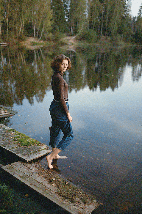 A woman stands barefoot on a wooden dock with her feet in the water, looking back at the viewer.