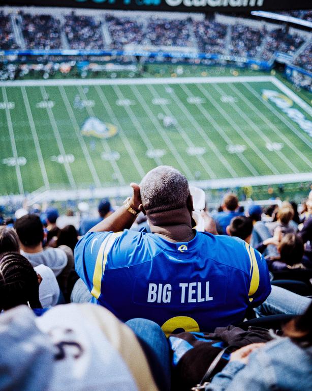 A man wearing a blue jersey with "BIG TELL" written on the back watches a football game from the stands.