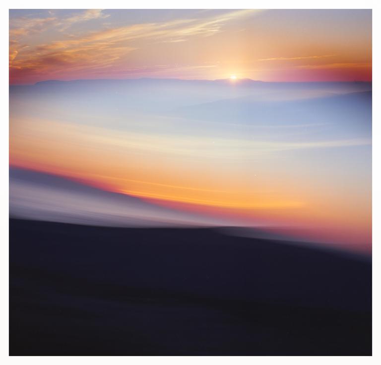 The sun sets over a hazy landscape with a dark foreground and a colorful sky.