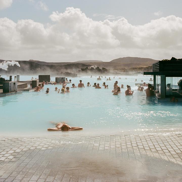 Many people are swimming in a light blue geothermal pool with steam rising from the water and mountains in the background.