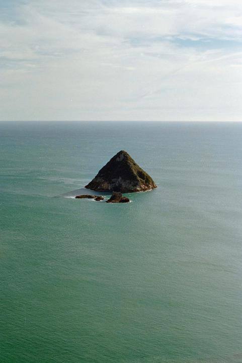A small, pointy island covered in green foliage rises from the ocean under a cloudy sky.