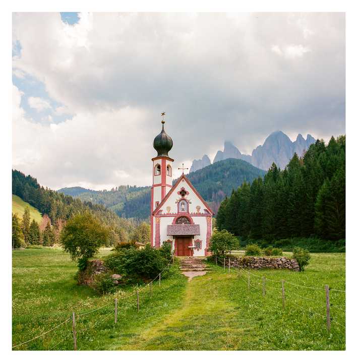A small church with a red and white exterior and a domed bell tower stands in a green meadow with mountains and a forest in the background.