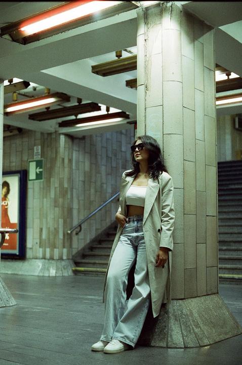 A woman wearing sunglasses, a crop top, and a trench coat leans against a pillar in a subway station.