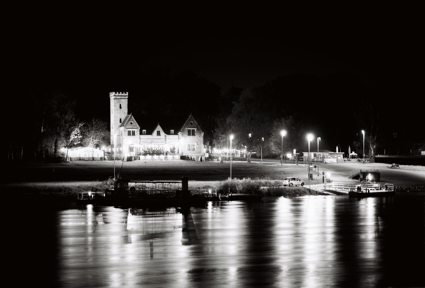 A large building with a tower and other structures are illuminated at night next to a river with reflections of light.