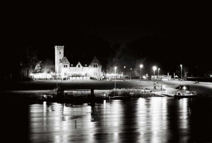 A large building with a tower and other structures are illuminated at night next to a river with reflections of light.