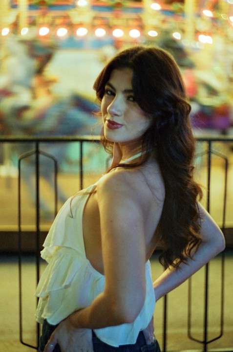 A woman with long, dark hair looks over her shoulder at the viewer, with a blurred background of colorful lights.