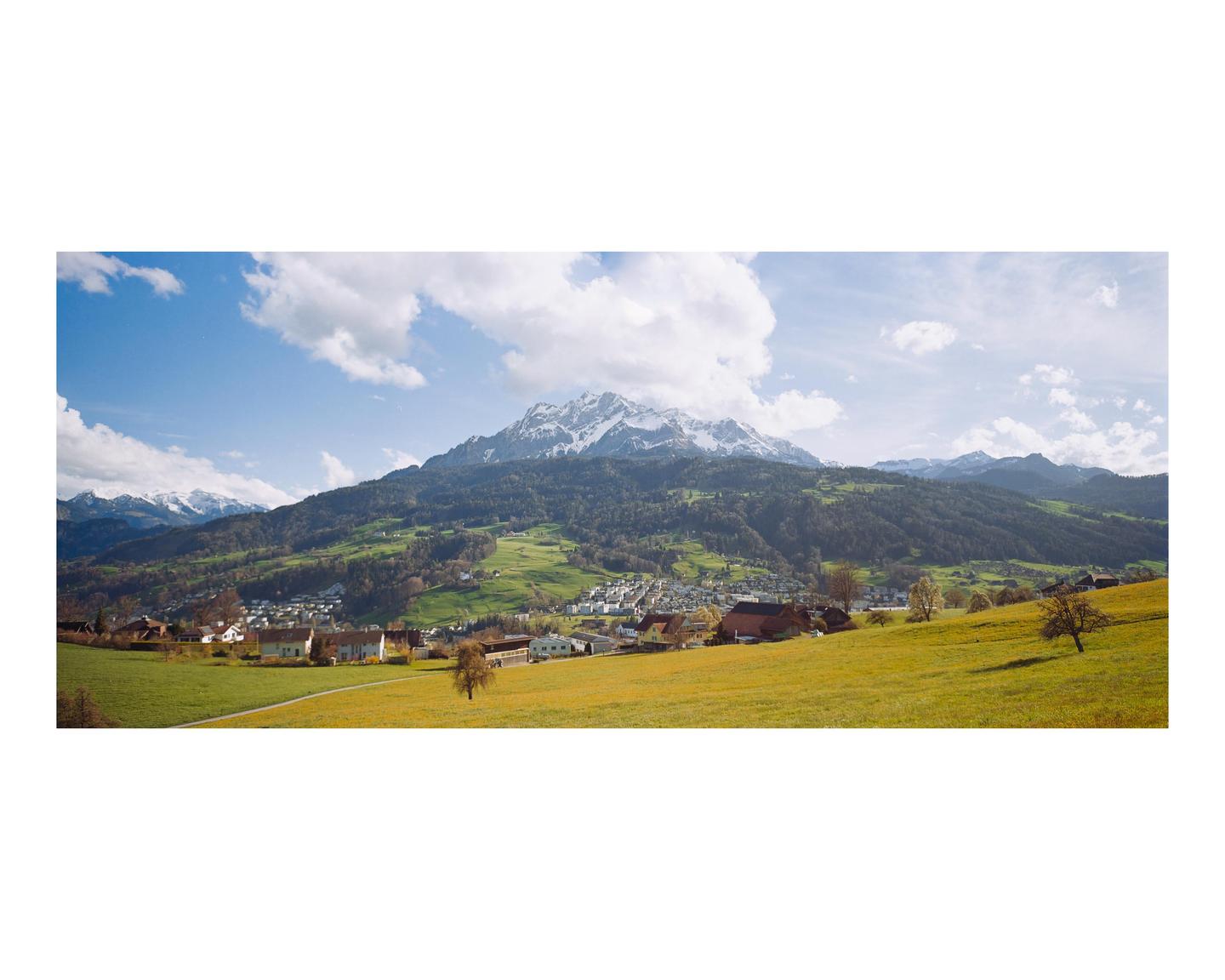 A panoramic landscape shows a village nestled in a valley with green hills and snow-capped mountains under a blue sky with clouds.