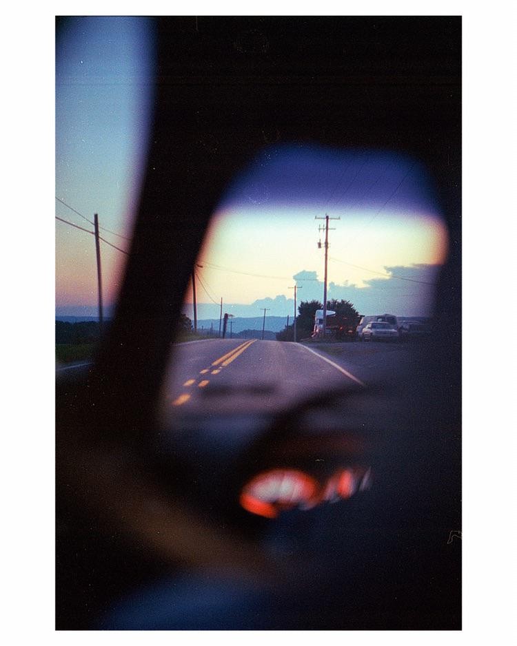 A road stretches into the distance, viewed through a car's rear window and side mirror at dusk.