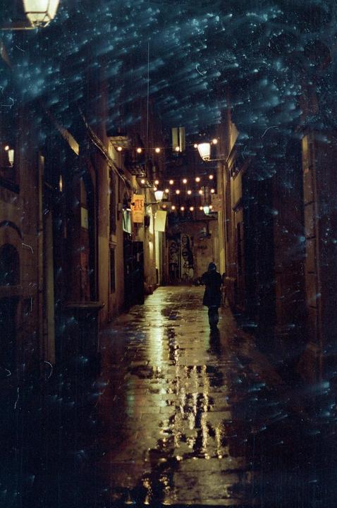 A person walks down a narrow, wet street at night, illuminated by string lights and street lamps.