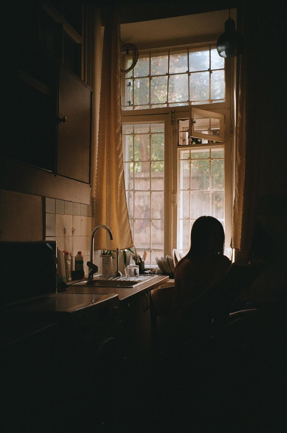 A woman sits at a kitchen counter in front of a sunlit window.