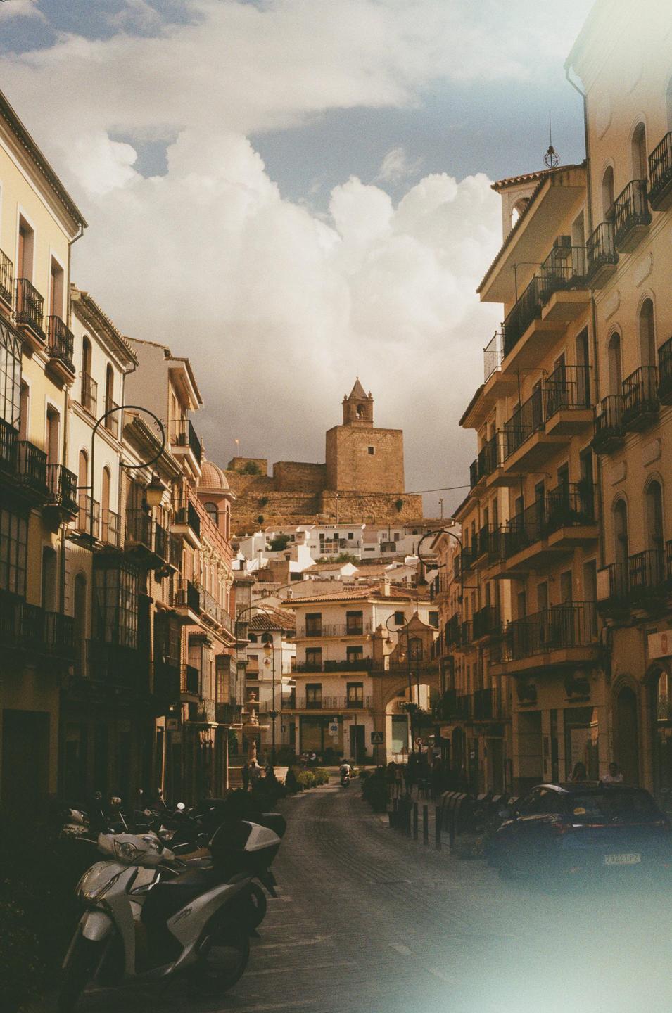 A narrow street in a Spanish town leads up to a historic building with a tower on a hill.