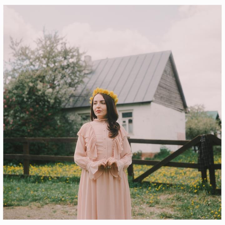 A woman wearing a flower crown and a long dress stands in a field with a house in the background.