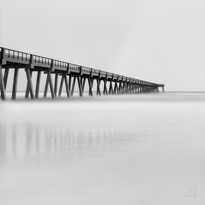 A long pier extends into the distance over calm water under a bright sky.