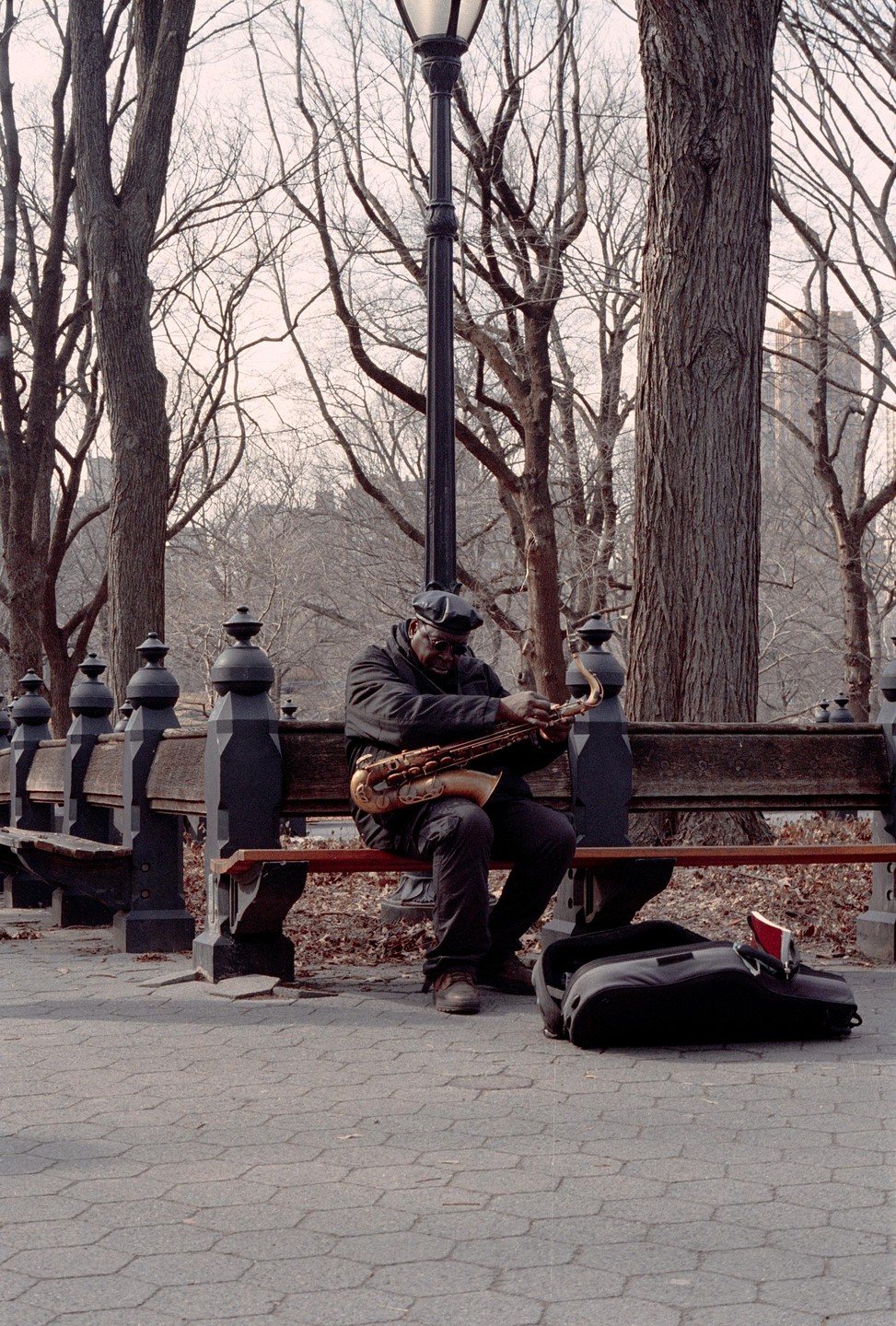 A man sits on a park bench, holding a saxophone, with a saxophone case on the ground beside him, surrounded by bare trees and a lamppost.
