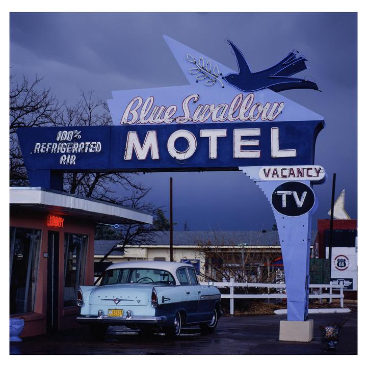 A vintage car is parked in front of the Blue Swallow Motel sign under a dark sky.