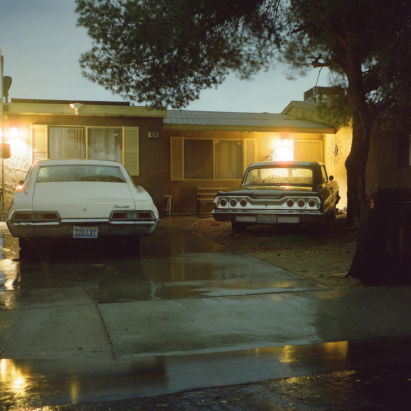 Two vintage cars are parked in front of a house at night, with wet pavement reflecting the light.
