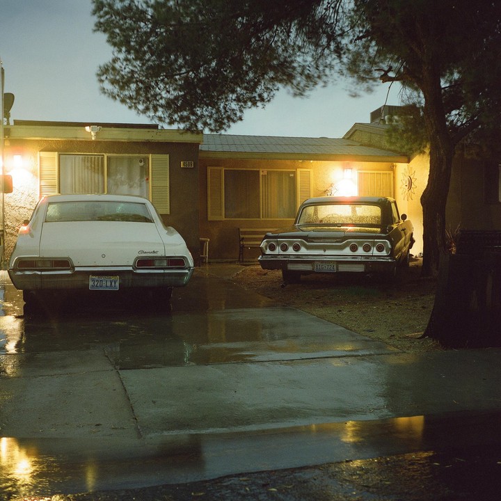 Two vintage cars are parked in front of a house at night, with wet pavement reflecting the light.