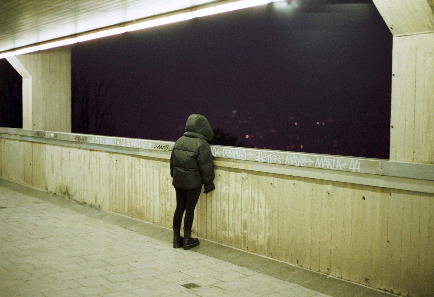 A person in a hooded jacket stands on a concrete walkway at night, looking out over a dark expanse with distant lights.