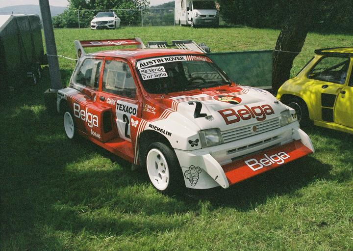 A red and white Austin Rover rally car is parked on a grassy field next to a yellow car.