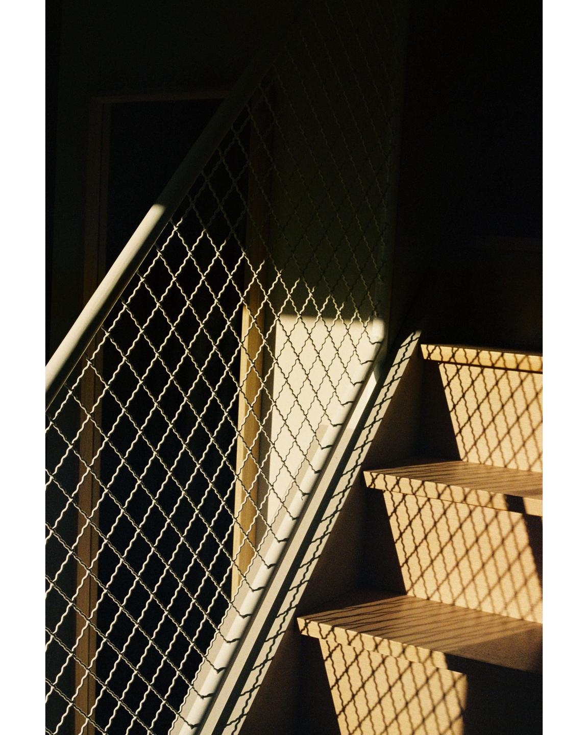 Sunlight casts a diamond pattern shadow from a metal railing onto a wooden staircase.