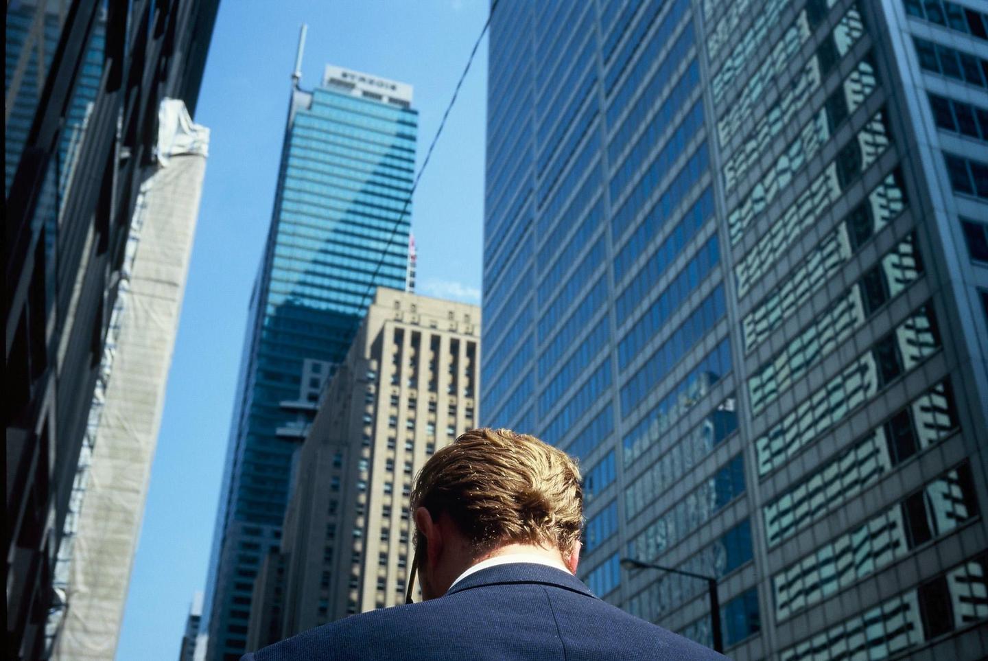 A man in a suit stands looking up at tall buildings in a city.