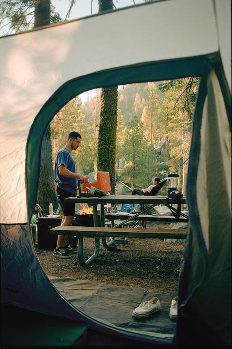 A man stands by a picnic table with a campfire in the background, viewed from inside a tent.